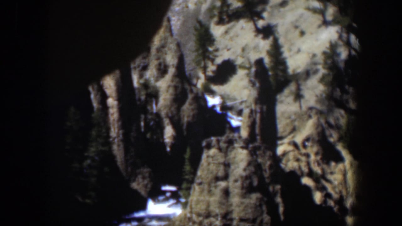 1951 Wyoming home-movie frame showing a steep rocky canyon wall, evergreens, white snow patches, and glinting water below.