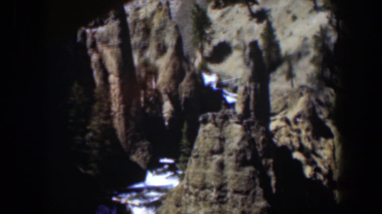 1951 8mm Wyoming landscape with vertical rock formations, scattered evergreens, melting snow, and bright water at the base.