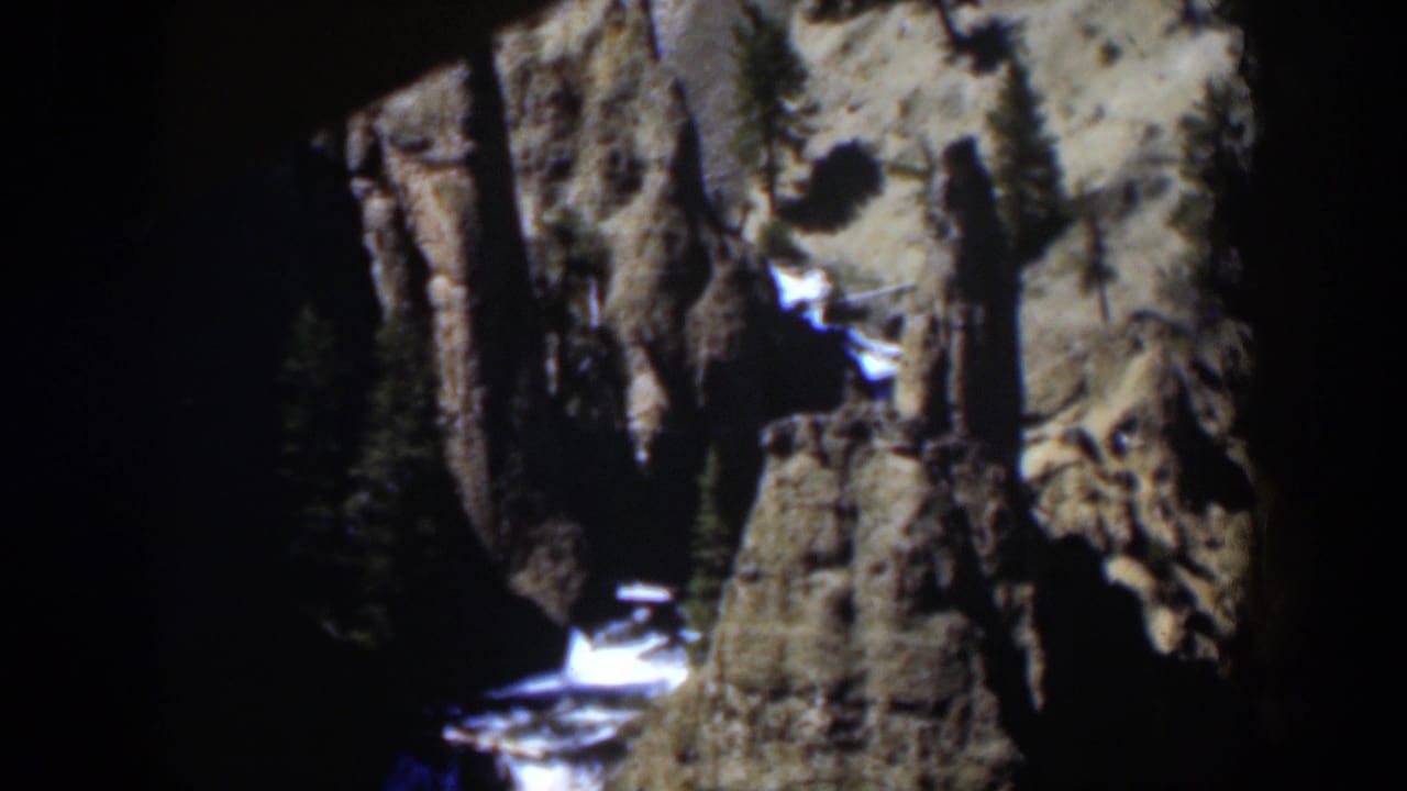 1951 Wyoming 8mm frame of shadowed canyon rock, pale sunlit stone, evergreen trees, snow patches, and bright water below.