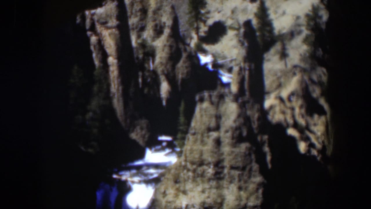 Waterfall dropping through a shadowed rocky canyon with evergreen trees nearby.