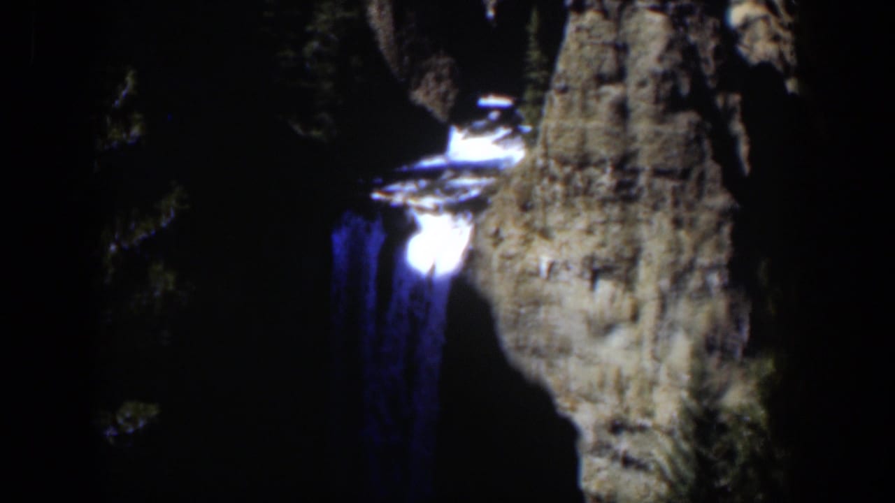 A blue-toned waterfall descends next to a tall rock outcrop in grainy 8mm footage from Wyoming in 1951.