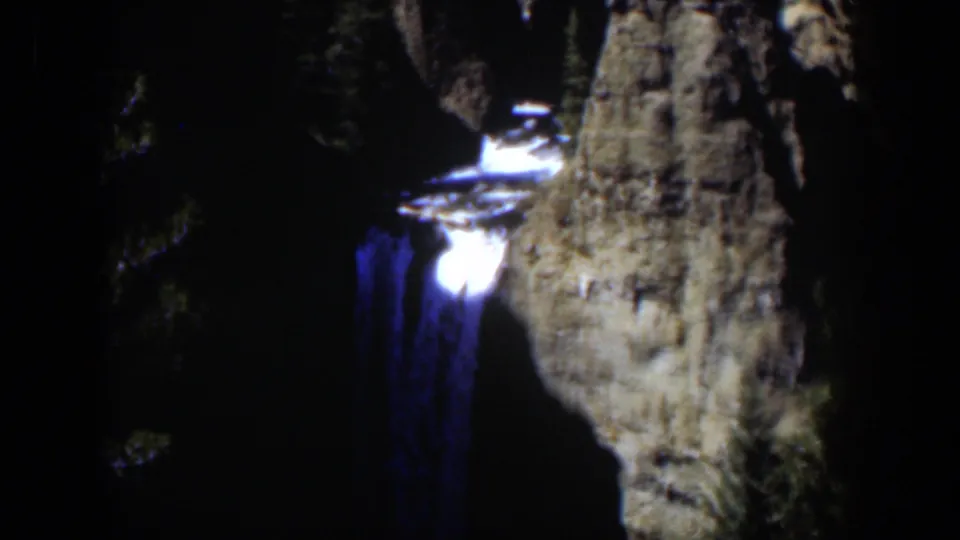A blue-toned waterfall descends next to a tall rock outcrop in grainy 8mm footage from Wyoming in 1951.