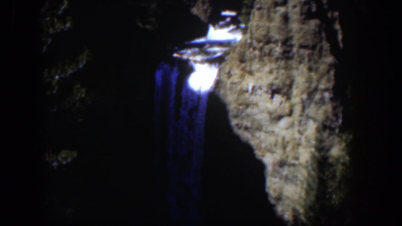 1951 Wyoming 8mm frame showing a blue-toned waterfall dropping beside a pale rugged cliff face in deep shadow.