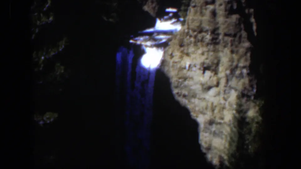 1951 Wyoming 8mm frame showing a blue-toned waterfall dropping beside a pale rugged cliff face in deep shadow.