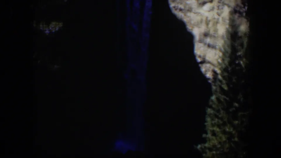 Wyoming home-movie frame showing a blue-toned waterfall descending through shadow beside jagged pale rock.