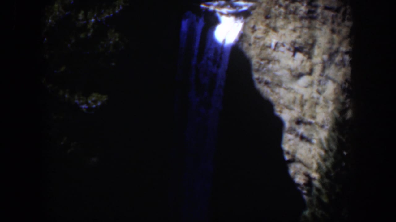1951 Wyoming 8mm frame with a luminous spot and blue trail near a dark canyon wall, with trees fading into shadow.