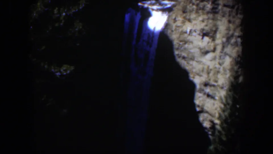 1951 Wyoming 8mm frame with a luminous spot and blue trail near a dark canyon wall, with trees fading into shadow.