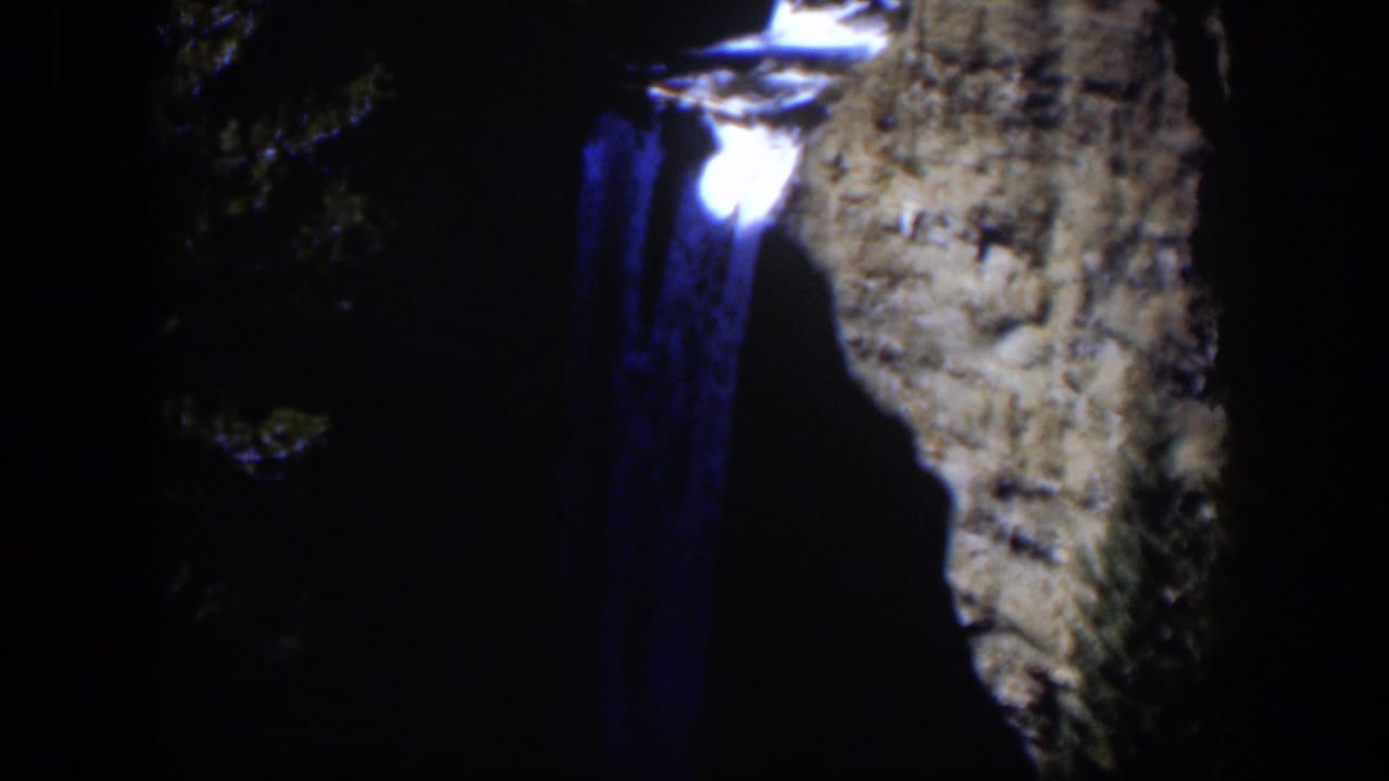 1951 Wyoming 8mm frame showing a glowing point and blue smear beside a shadowed rocky slope and foliage.