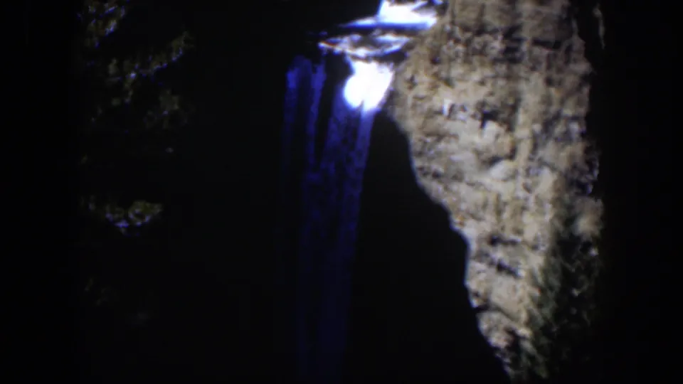 1951 Wyoming 8mm frame showing a glowing point and blue smear beside a shadowed rocky slope and foliage.