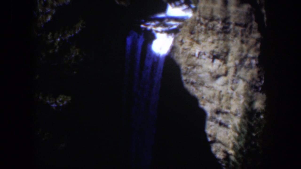 Blue-lit waterfall beside a rugged cliff face in deep night shadow.