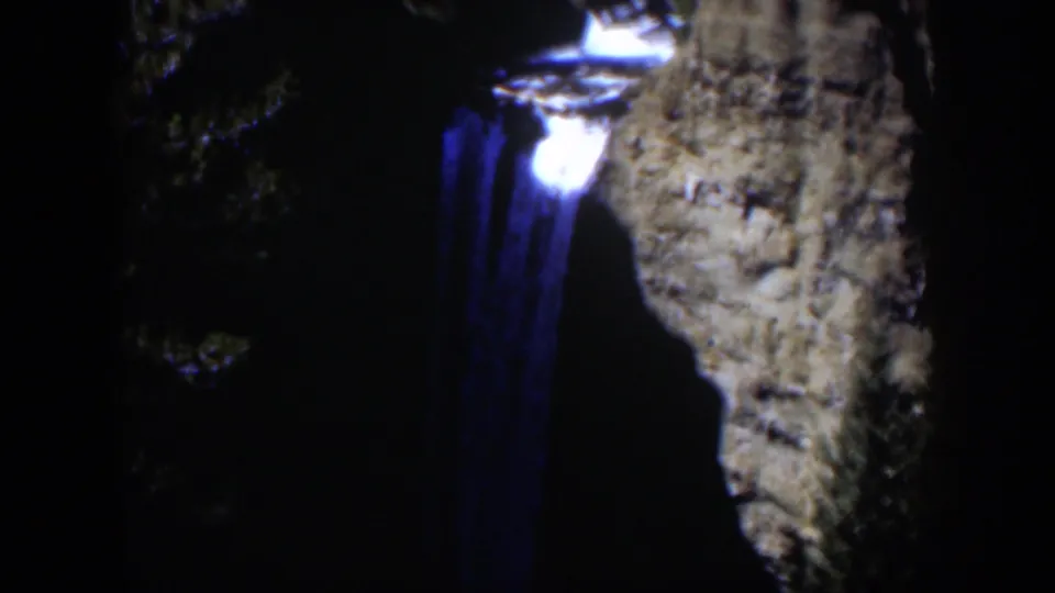 1951 Wyoming 8mm frame showing a blue illuminated waterfall dropping beside a rough cliff face in dark forest surroundings.