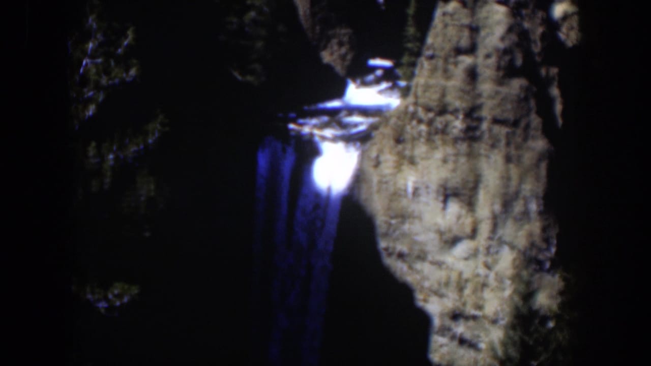 1951 color 8mm frame of the blue-lit waterfall, with a bright lip of water and a steep cliff face visible through low-light grain.
