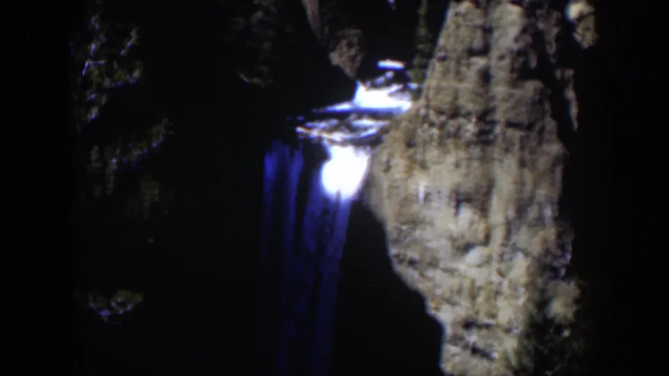 1951 color 8mm frame of the blue-lit waterfall, with a bright lip of water and a steep cliff face visible through low-light grain.