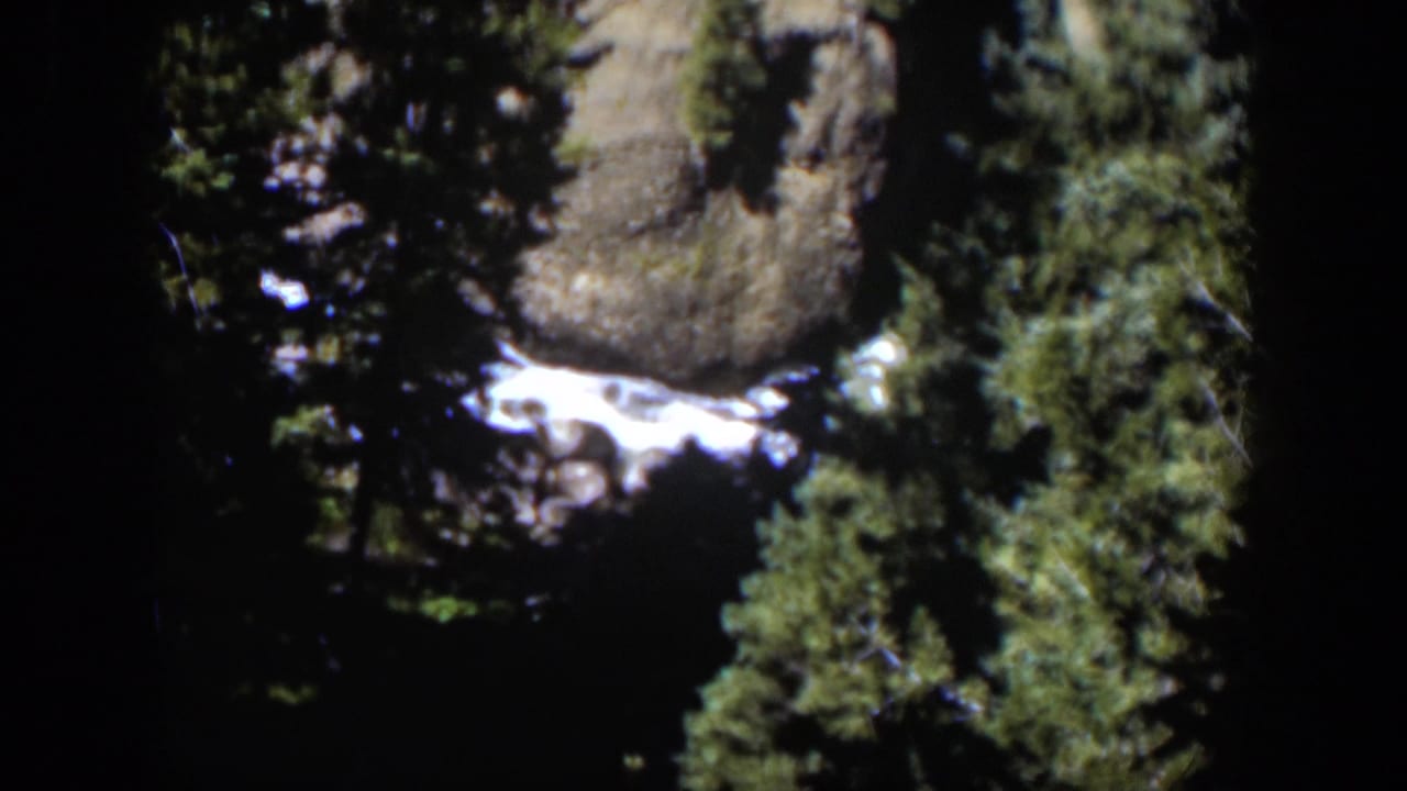 1951 Wyoming home-movie frame showing bright water, rugged rocks, and dense green trees in a mountain setting.
