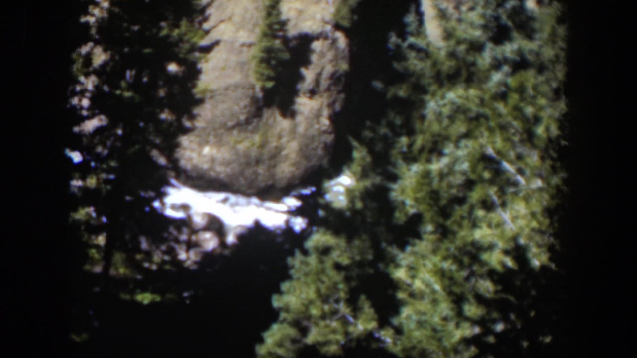 Vintage 8mm Wyoming frame with rushing white water at the base of rocks, framed by dark trees and green foliage.