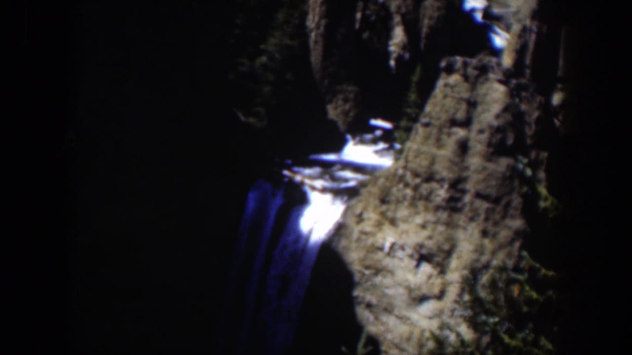 1951 Wyoming frame showing a narrow waterfall descending past a rock face with trees in heavy shadow.