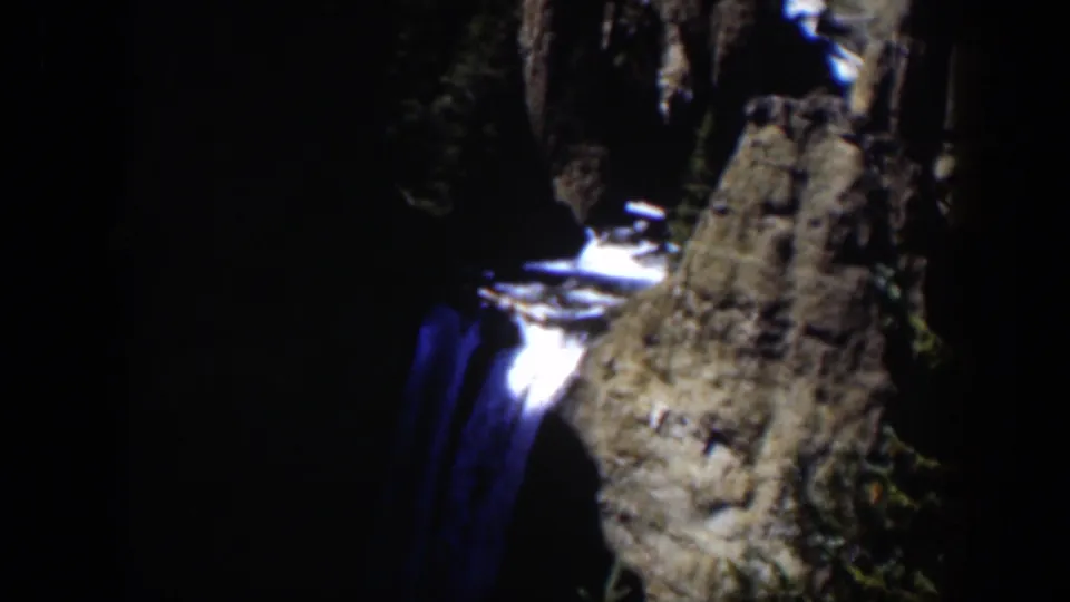 1951 Wyoming frame showing a narrow waterfall descending past a rock face with trees in heavy shadow.