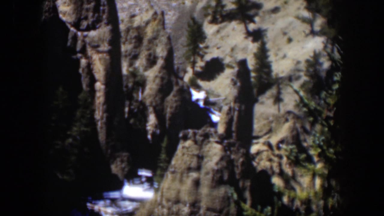 Rocky canyon wall with evergreens and a bright stream below dark shadow.