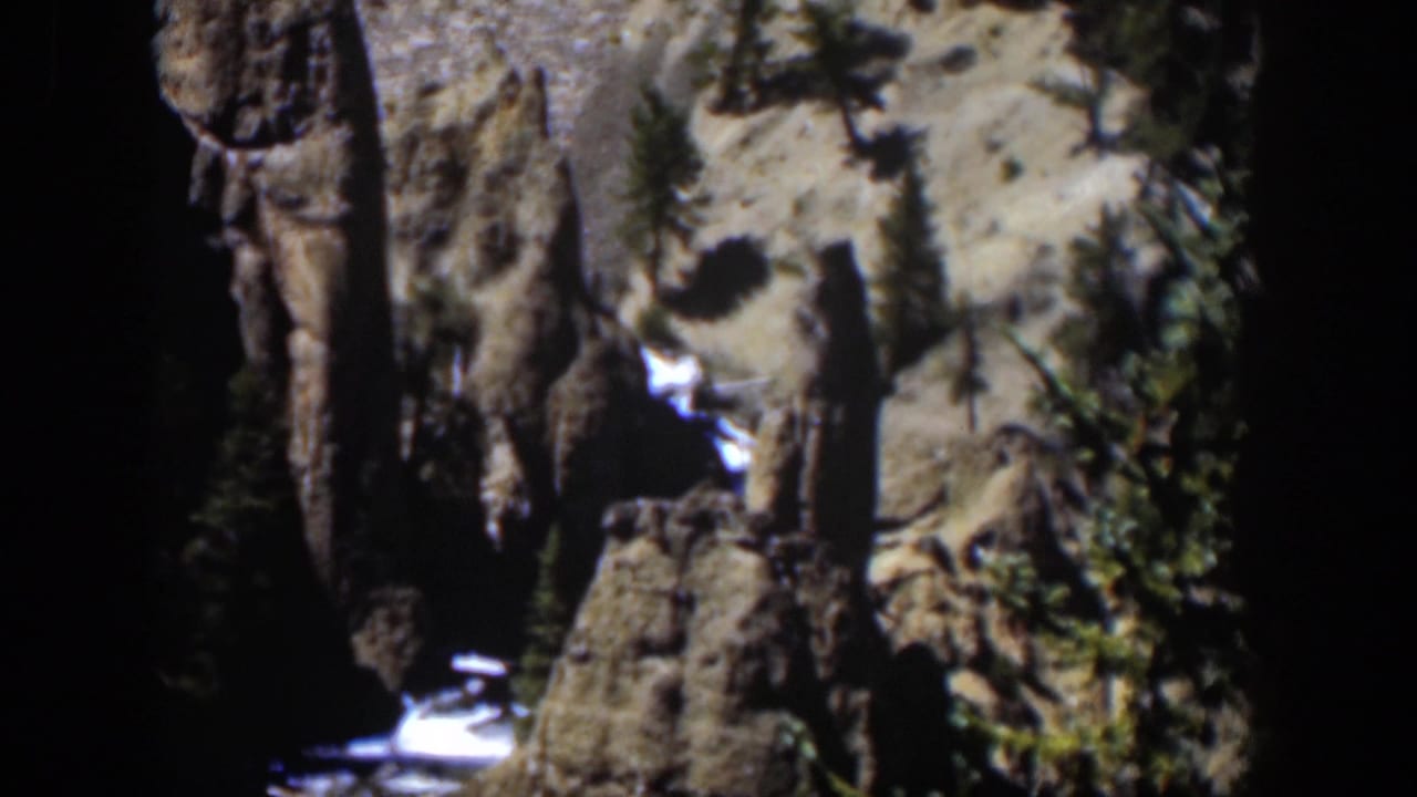 1951 Wyoming 8mm frame with rugged vertical rock, scattered trees, pale sunlit ground, and a small bright stream below.
