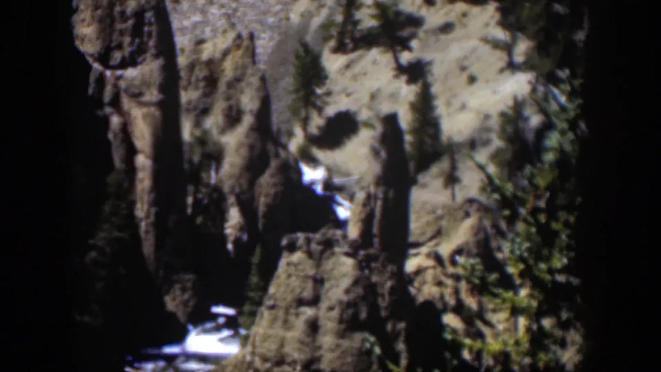 1951 Wyoming 8mm frame with rugged vertical rock, scattered trees, pale sunlit ground, and a small bright stream below.