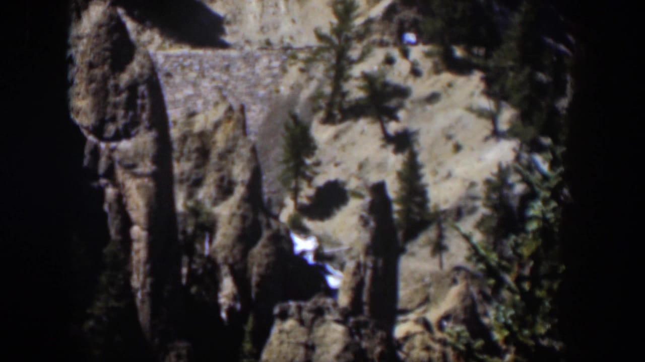 1951 Wyoming 8mm frame showing a tall rock face, dark ravine, evergreen trees, and a bright stream through the canyon floor.