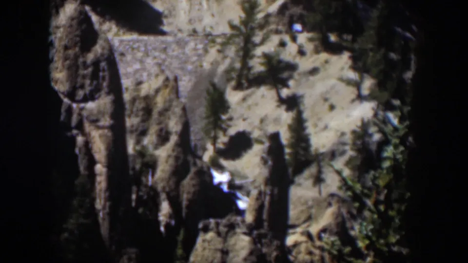 1951 Wyoming 8mm frame showing a tall rock face, dark ravine, evergreen trees, and a bright stream through the canyon floor.