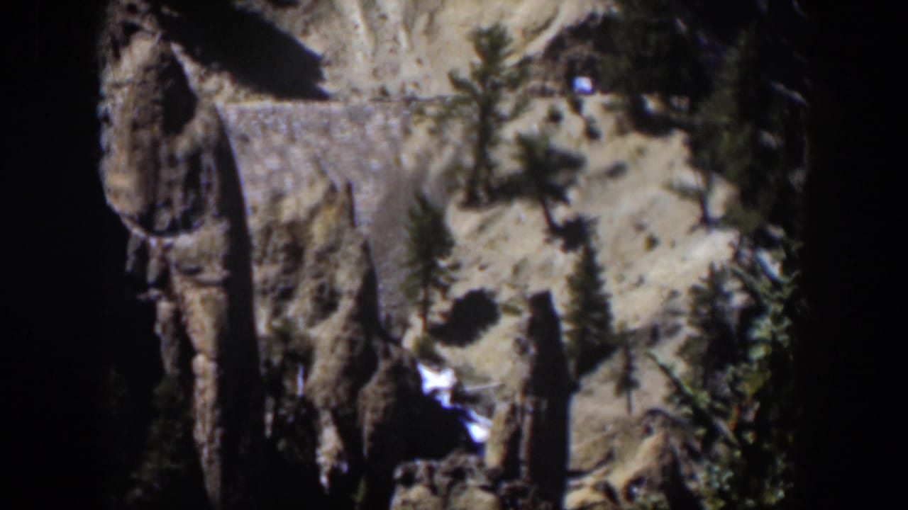 1951 Wyoming 8mm frame of rocky canyon terrain, evergreen growth, pale sunlit slope, and white water near the lower edge.