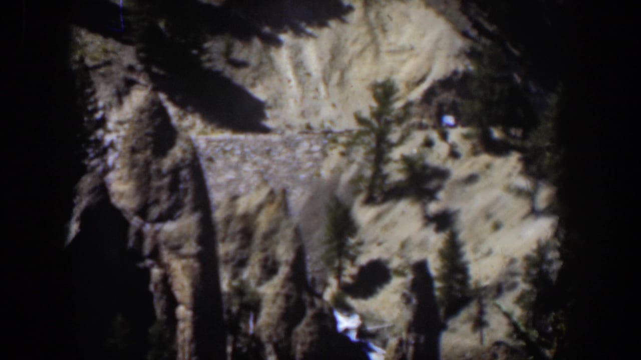 1951 Wyoming 8mm view of pale canyon terrain, dark rock formations, scattered evergreens, and a stone retaining wall.