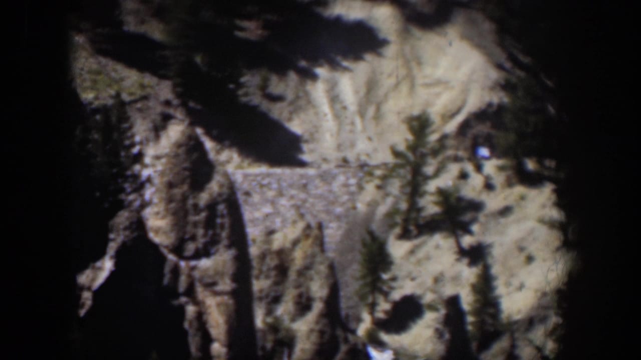 1951 Wyoming home-movie frame of a rugged canyon slope with a stone wall, evergreen trees, and high-contrast sunlight.
