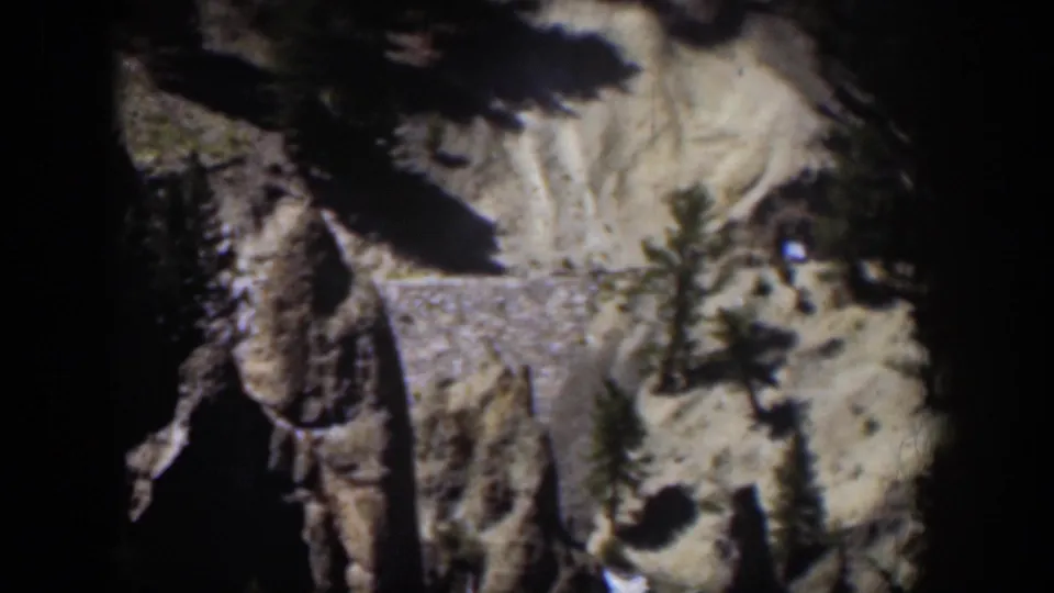 1951 Wyoming home-movie frame of a rugged canyon slope with a stone wall, evergreen trees, and high-contrast sunlight.