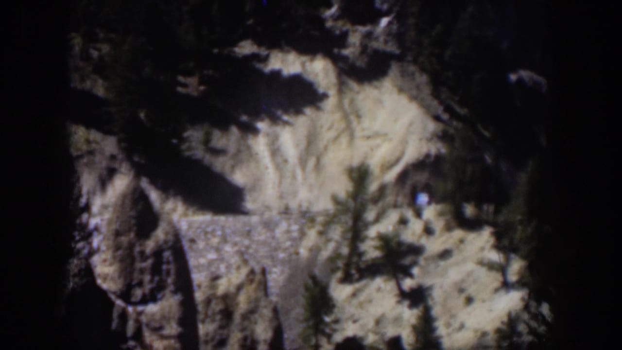 1951 Wyoming 8mm frame showing a dry canyon hillside, dark rock shapes, scattered evergreens, and a low stone wall.