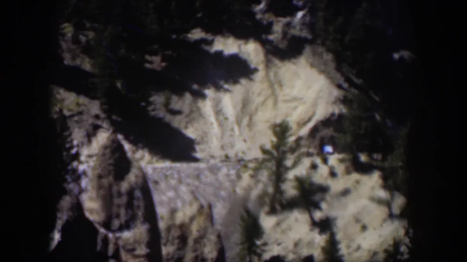 1951 Wyoming 8mm frame showing a rocky mountain slope, evergreen trees, dark shadows, and pale snow or exposed ground.