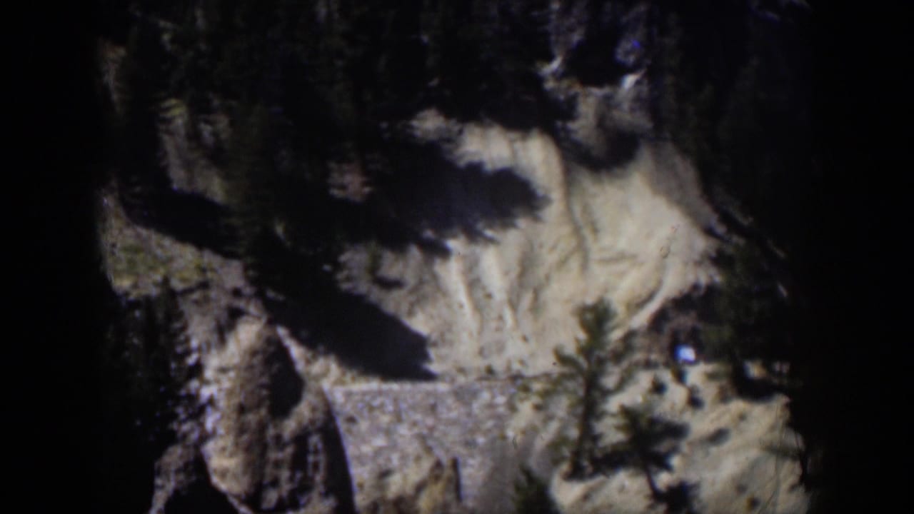 1951 Wyoming 8mm frame of a rugged slope with trees, shadowed rock, pale ground, and a small bright spot near the right edge.