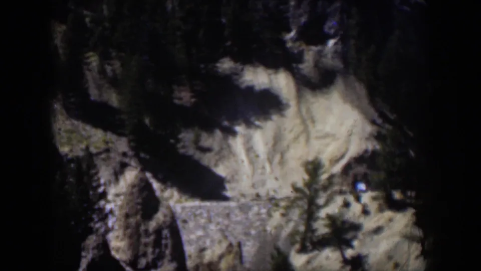 1951 Wyoming 8mm frame of a rugged slope with trees, shadowed rock, pale ground, and a small bright spot near the right edge.