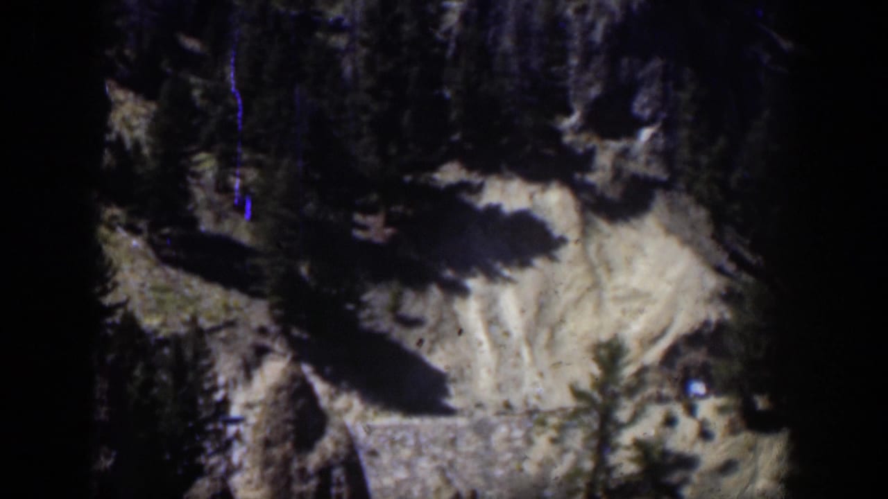 1951 Wyoming 8mm frame with a darker view of the rocky mountain slope, evergreen forms, pale ground, and a blue vertical film mark.