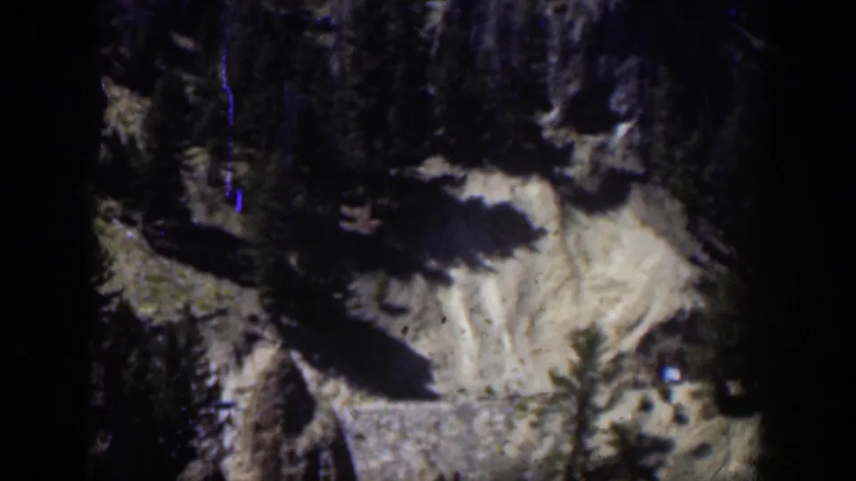 1951 Wyoming 8mm frame with a darker view of the rocky mountain slope, evergreen forms, pale ground, and a blue vertical film mark.