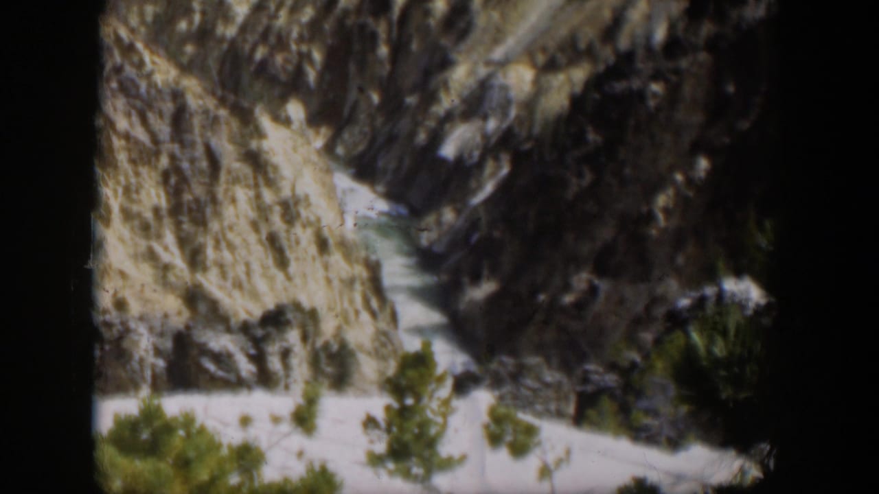 1951 Wyoming home-movie frame with a rushing stream cutting through steep rock walls and sparse evergreen growth.
