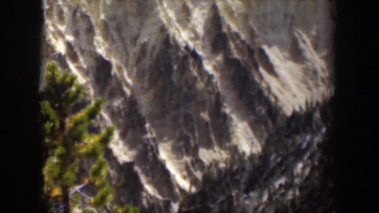 1951 Wyoming 8mm frame holding on a pine tree against a rugged, snow-marked mountain face with visible film-edge vignetting.