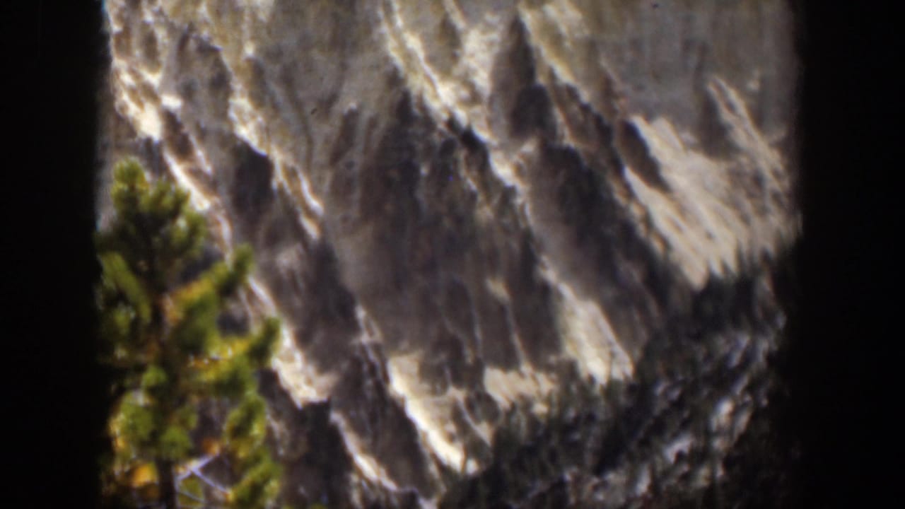 Rocky mountain face with a foreground evergreen and dark film edges.
