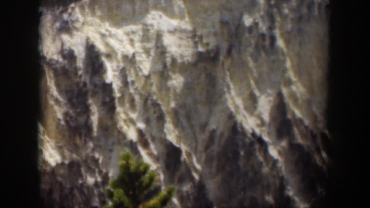 1951 Wyoming 8mm frame showing a distant pale mountain face, scattered snow in crevices, and a green evergreen near the bottom.