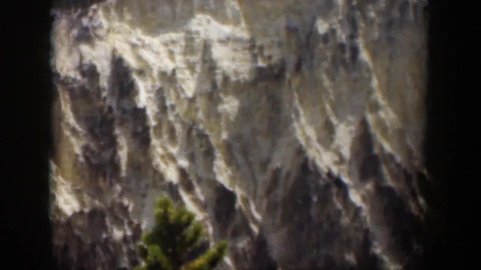 1951 Wyoming 8mm frame showing a distant pale mountain face, scattered snow in crevices, and a green evergreen near the bottom.