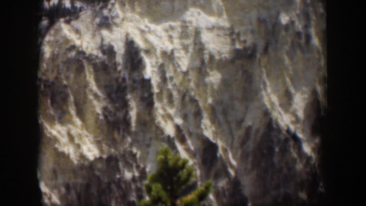 1951 Wyoming scenic frame of a snow-marked rocky mountain slope, dark shadowed ridges, and a green evergreen in the foreground.