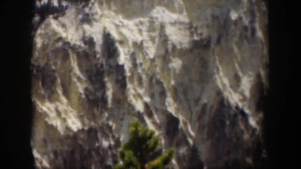 1951 Wyoming scenic frame of a snow-marked rocky mountain slope, dark shadowed ridges, and a green evergreen in the foreground.