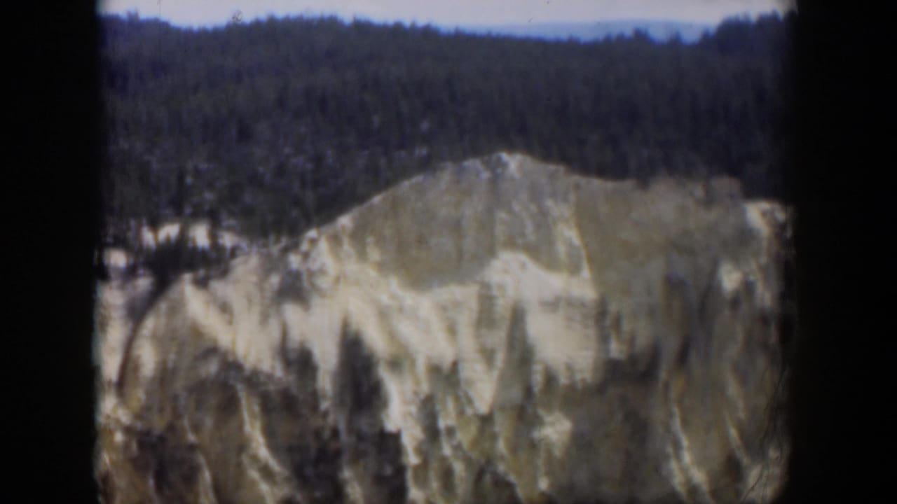 1951 Wyoming 8mm frame showing a pale eroded mountain face beneath dense evergreen forest and a hazy sky.