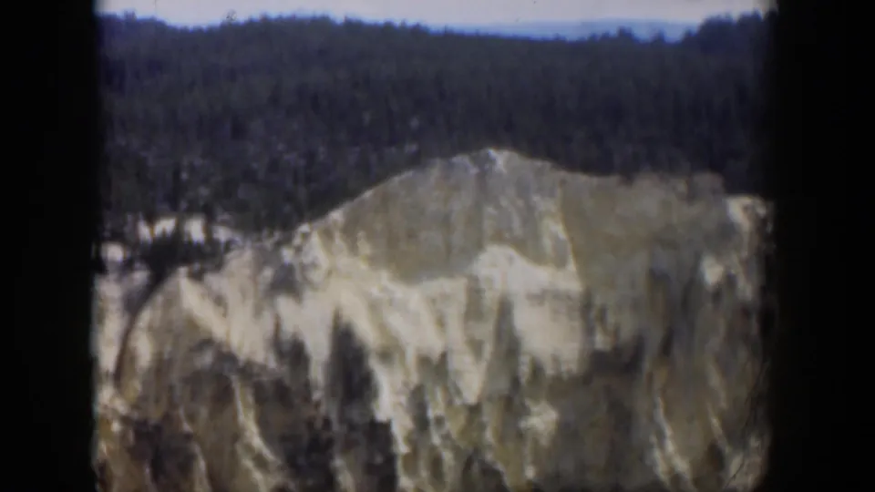 1951 Wyoming 8mm frame showing a pale eroded mountain face beneath dense evergreen forest and a hazy sky.