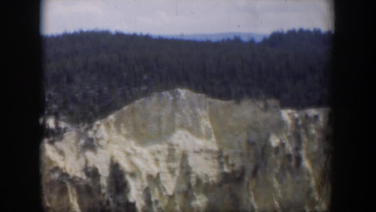 1951 Wyoming 8mm view of a light-colored cliff or slope backed by dense trees, framed by dark film-scan edges.