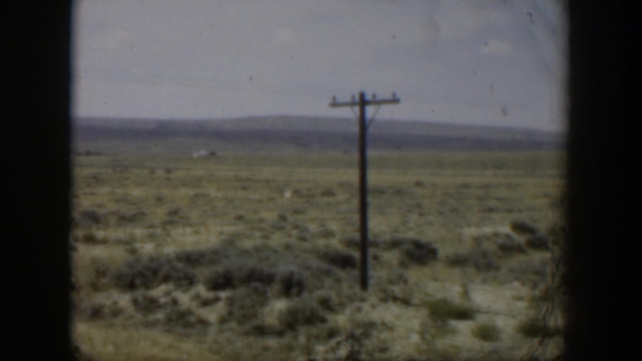 1951 Wyoming 8mm frame showing scrubland, a solitary utility pole, pale sky, and distant low mountains from a moving viewpoint.
