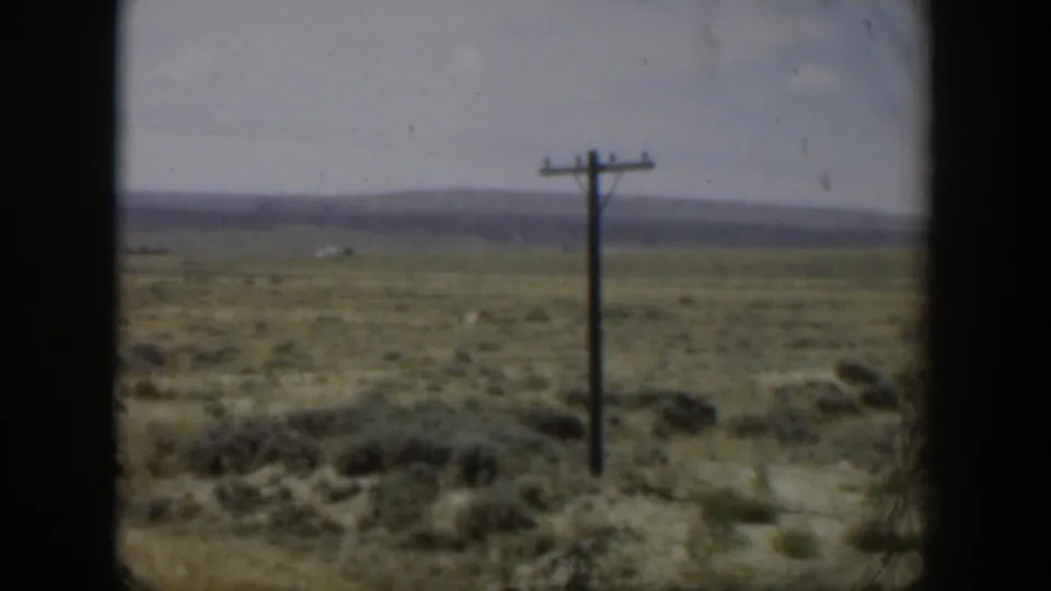 1951 Wyoming 8mm frame showing scrubland, a solitary utility pole, pale sky, and distant low mountains from a moving viewpoint.