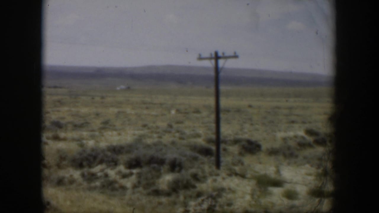 1951 Wyoming roadside view with a dark film edge, wooden pole, brushy field, and low mountains across the horizon.