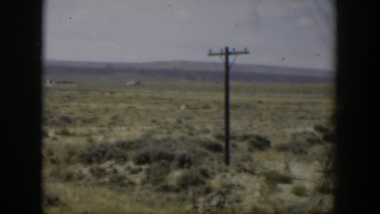 1951 Wyoming landscape frame showing a utility pole, sparse roadside brush, open field, and low mountains in soft 8mm color.